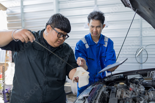 Two mechanics examine a car engine together. One holds a clipboard, the other a laptop. They focus on the engine's components closely. Their expressions show concentration and teamwork