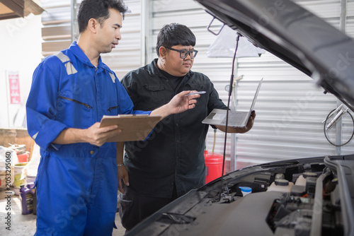 Two mechanics examine a car engine together. One holds a clipboard, the other a laptop. They focus on the engine's components closely. Their expressions show concentration and teamwork