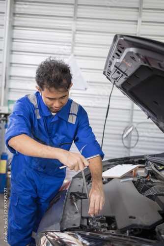 Two mechanics examine a car engine together. One holds a clipboard, the other a laptop. They focus on the engine's components closely. Their expressions show concentration and teamwork