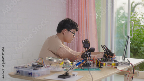 Asian young student using a laptop at home while working on a robotics project with electronic components and robotic devices. Concept of STEM education, coding, and technology learning.