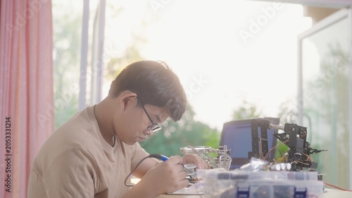 Asian young student using a soldering iron for electronics and robotics projects at home with robotic equipment and a laptop. Concept of STEM education, engineering skills, and innovation.