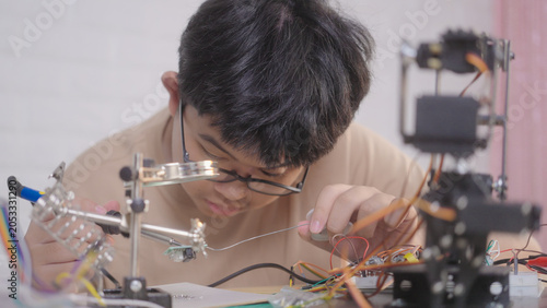 Asian young student using a soldering iron for electronics and robotics projects at home with robotic equipment and a laptop. Concept of STEM education, engineering skills, and innovation.