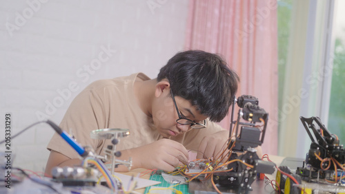 Asian young student wiring a robotics project at home with robotic devices, electronics, and a laptop. Concept of STEM education, engineering skills, innovation, and hands-on technology learning.