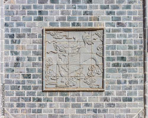 Traditional Chinese Bas-Relief Dragon Wall Sculpture. Stone carving of a mythical dragon amidst clouds on a central panel within a rustic, weathered gray brick wall in Asian architectural style.