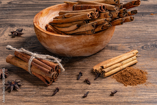 Cinnamon sticks in wooden bowl with ground cinnamon powder and various spices scattered on a rustic surface