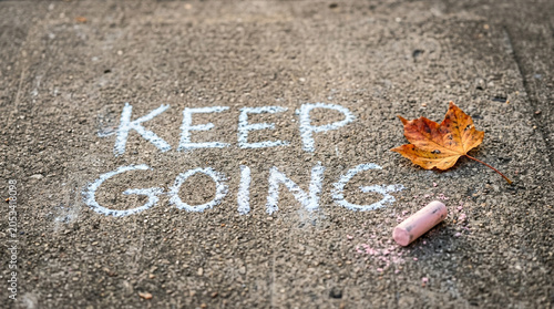 Inspirational Keep Going message written in chalk on asphalt, motivational quote on sidewalk with autumn leaf, perseverance concept on urban pavement