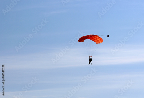 Fotografie Minimalist horizontal shot of a lone skydiver under a vibrant orange parachute canopy drifting through a vast, calm light blue sky with plenty of copy space for text