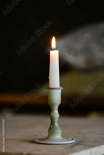 A single white lit candle in a ceramic holder casting a warm glow against a dark, rustic background.