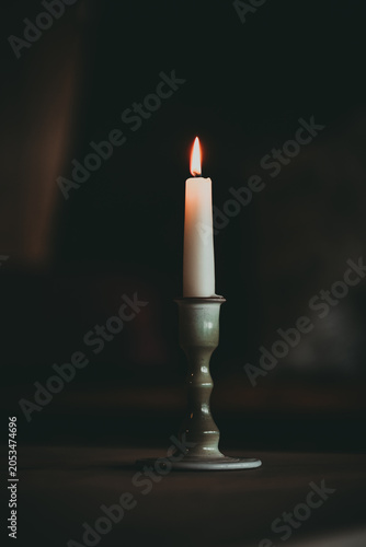 A single white lit candle in a ceramic holder casting a warm glow against a dark, rustic background.