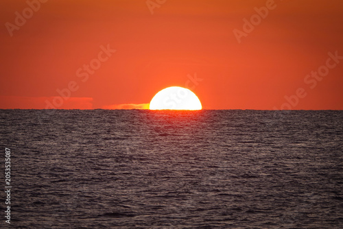 Obraz na plátně LENS FLARE: Sun sinks halfway below the horizon, turning the sky a deep orange