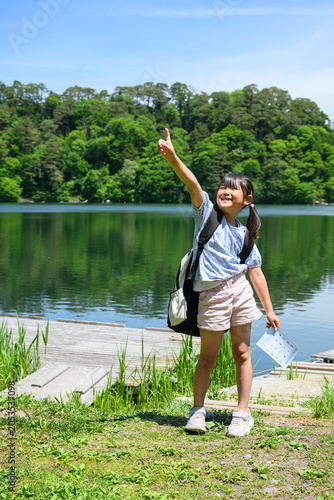 新緑の湖畔でしおりを持ち元気に手を挙げる遠足の女子小学生