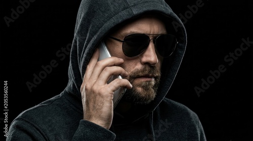 A mysterious man in a dark hoodie and sunglasses making a phone call to impersonate a trusted source.