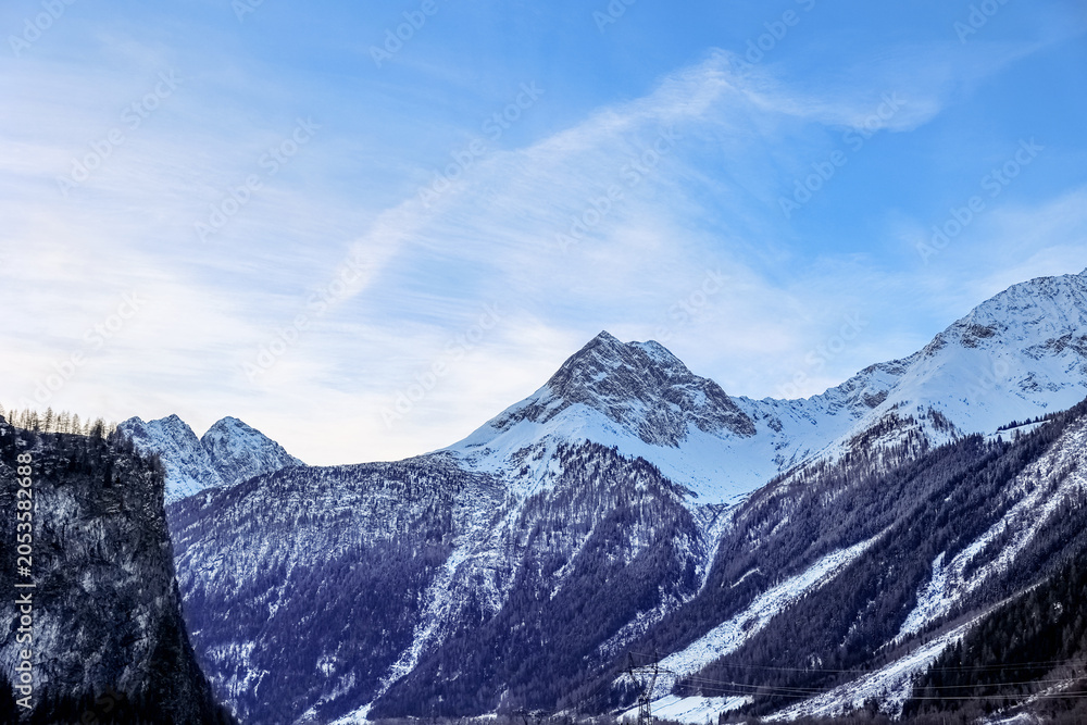 Obraz premium Snowy mountain peaks and alpine forest landscape under blue sky with wispy clouds, winter nature scenery, the winter nature scenery features rugged rock formations and snow-covered trees