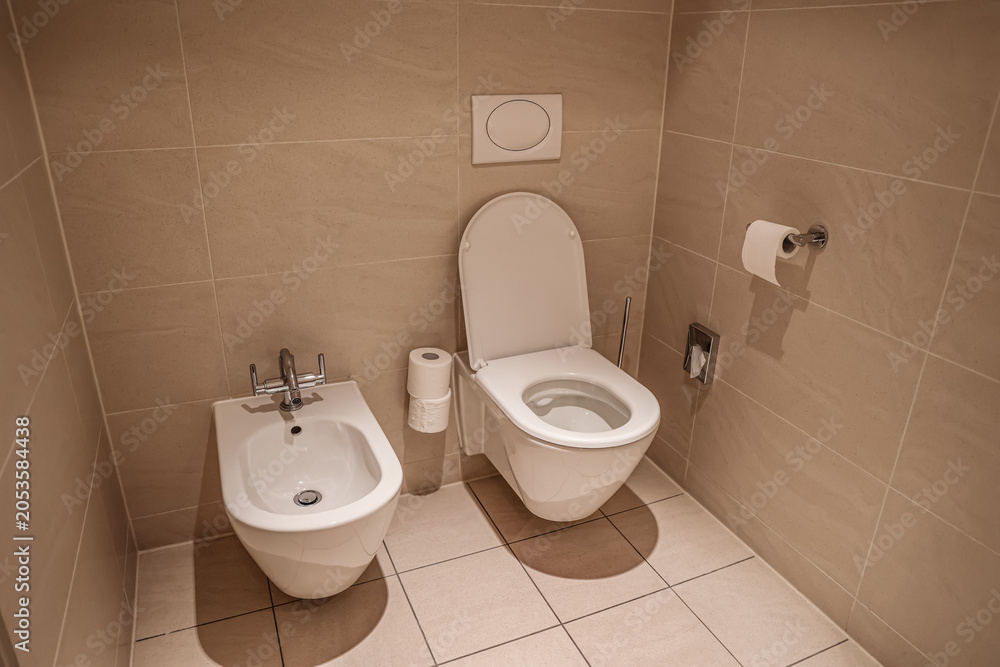 Fototapeta premium Modern bathroom interior with white ceramic toilet and bidet, beige tiled walls and floor, clean restroom design