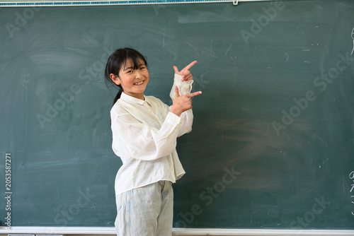 教室の黒板の前で笑顔で指差しをする高学年の女子小学生