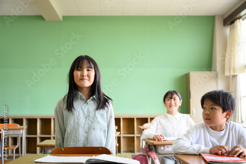 教室の席で起立して元気に発表する女子小学生と話を聞くクラスメイト