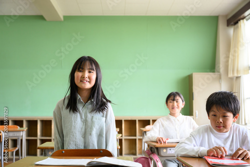 教室の席で起立して元気に発表する女子小学生と話を聞くクラスメイト