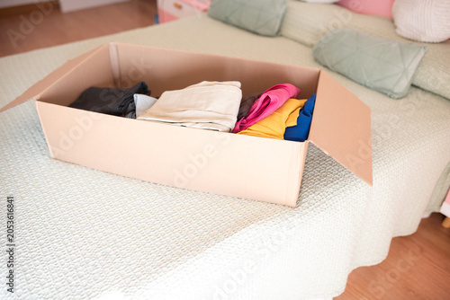 Bild auf Leinwand Open cardboard moving box with colorful folded clothes on a bed.