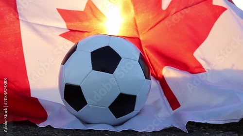 A traditional black and white soccer ball sits before a flowing Canadian flag. Bright sunlight shines through the flag, highlighting the upcoming 2026 World Cup hosted by Canada and USA