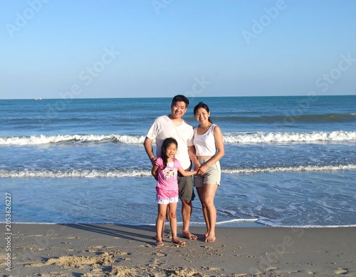 happy Asian family of three standing together on a sandy beach beside the ocean