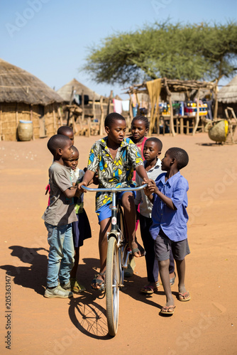 In a warm, rural african village, a group of children gathers around as one proudly rides a bicycle. Laughter and camaraderie fill the air under a bright blue sky. Generative AI.