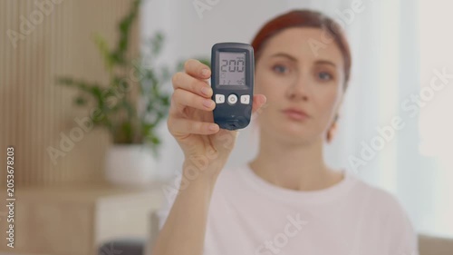 A woman holds up a glucometer that shows a high blood sugar level of 200 mg dL. She is in a bright indoor space with plants around her.