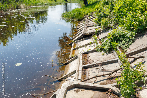 Damaged concrete reinforcement slabs collapsing along the bank of a calm river or canal surrounded by green vegetation.