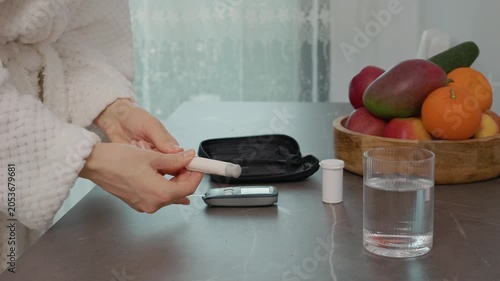 A woman in a kitchen checks her blood sugar level with a glucometer. She prepares to take a blood sample while managing diabetes and monitoring health.