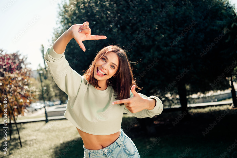 Naklejka premium Playful young woman in crop top and jeans framing a selfie with her hands in a sunny park, smiling and sticking out her tongue in a carefree outdoor lifestyle portrait