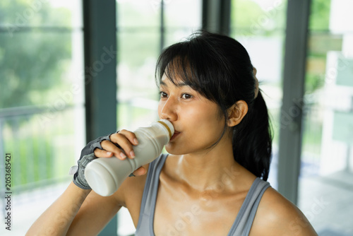 Asian woman in gray sportswear smiles at the camera while holding a bottle of protein drink in a bright gym with windows.
