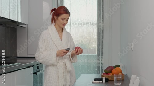 In a bright kitchen, a woman checks her blood sugar with a glucometer while holding an apple. She is focused on managing her health at home.