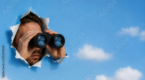 Creative conceptual image of a man looking through binoculars from behind torn blue paper with clouds in the background. Symbolic representation of vision, search, opportunity, curiosity, surveillance