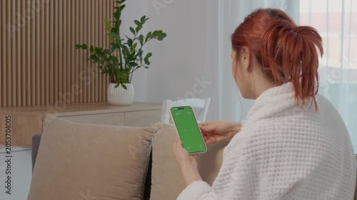 A woman sits on a sofa in her home and looks at a green screen phone. She interacts with it while relaxing in the living room during the day.