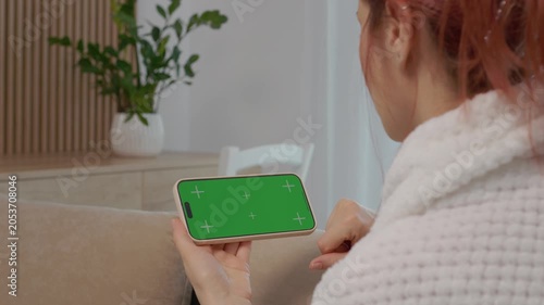 A woman sits on a sofa in her living room holding a phone with a green screen. She focuses her attention on the phone while relaxed at home.