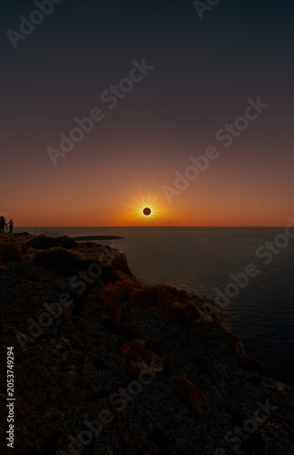 Silhouettes of people and a family with the father lifting his son to observe a total solar eclipse with a prominent starburst over the ocean coast of Menorca.