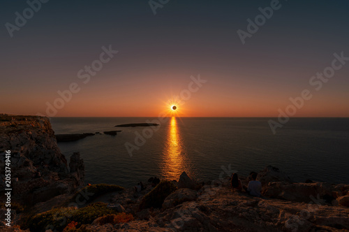 People watching a total solar eclipse at sunset from the rocky coast of Menorca with sunburst reflection on ocean