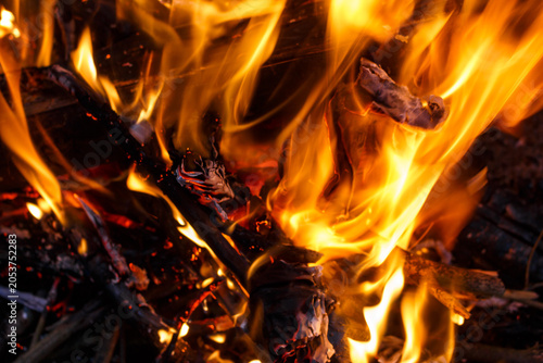 Vibrant orange and yellow flames dance fiercely around burning wood logs, creating a mesmerizing, abstract texture of heat and light in the darkness