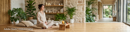 Serene woman meditating in a sunlit natural living room with plants and wooden floor for wellness background, mindfulness banner, and calm interior design