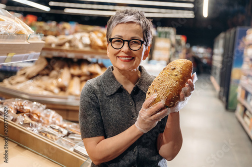 Fotografie Mature Asian woman with short gray hair wearing glasses holds a loaf of bread in
