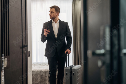 Konstfotografi Businessman in formal suit holding smartphone and suitcase, standing in hotel ro