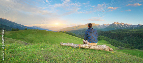 Man traveler sitting on a wooden log and looking at a beautiful sunset over mountain valley. Young tourist enjoying scenic alpine nature landscape panorama