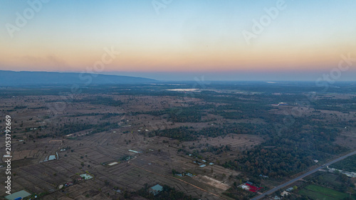 Fotografie An aerial view of a vast rural landscape at sunset, with warm orange and golden hues fading into a calm blue sky above farmland and scattered villages