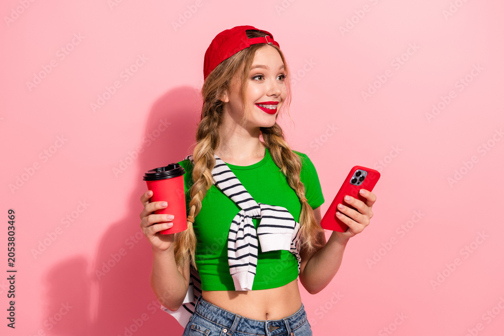 Fototapeta premium Young stylish girl with green top red cap holding coffee and phone against pink background looking happy and fashionable