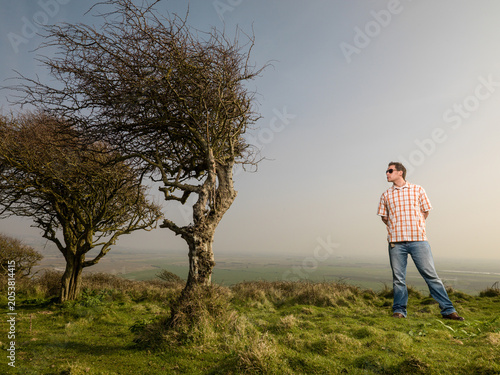 Konstfotografi Young man looking at wind blown tree on top of coastal hill.