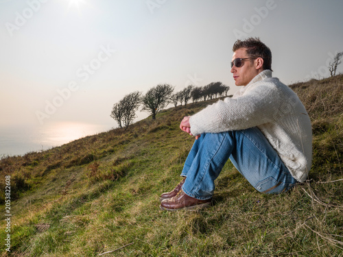 Konstfotografi Young man wearing sunglasses and thick wool jumper looking from hill out to sea