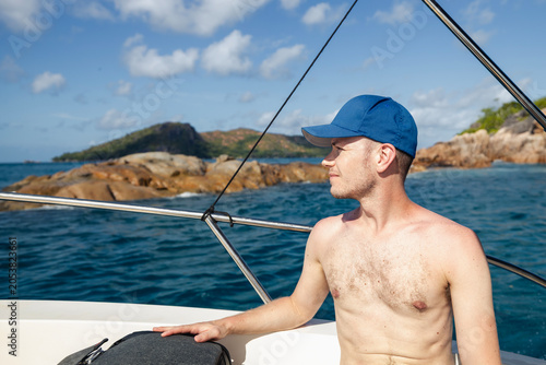 Fotografie Shirtless male tourist with cap sitting in speedboat cruising along tropical coastline of Praslin island in Seychelles