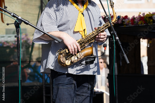 Male saxophonist wearing a checkered shirt and yellow scarf performs outdoors, holding a saxophone with microphones and a blurred audience in the background