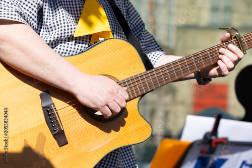 Close-up of a person playing an acoustic guitar outdoors, wearing a checkered shirt and yellow scarf, with music sheets visible in the background