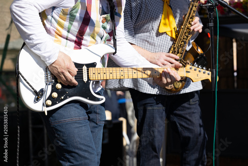 Male guitarist wearing a colorful striped shirt plays electric guitar on stage with a saxophonist in the background during a live music performance outdoors