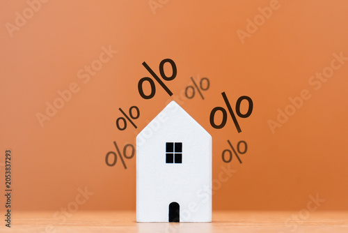 A white house model surrounded by floating percentage signs. Mortgage interest rates, property taxes, real estate investment returns, and housing market inflation concept.